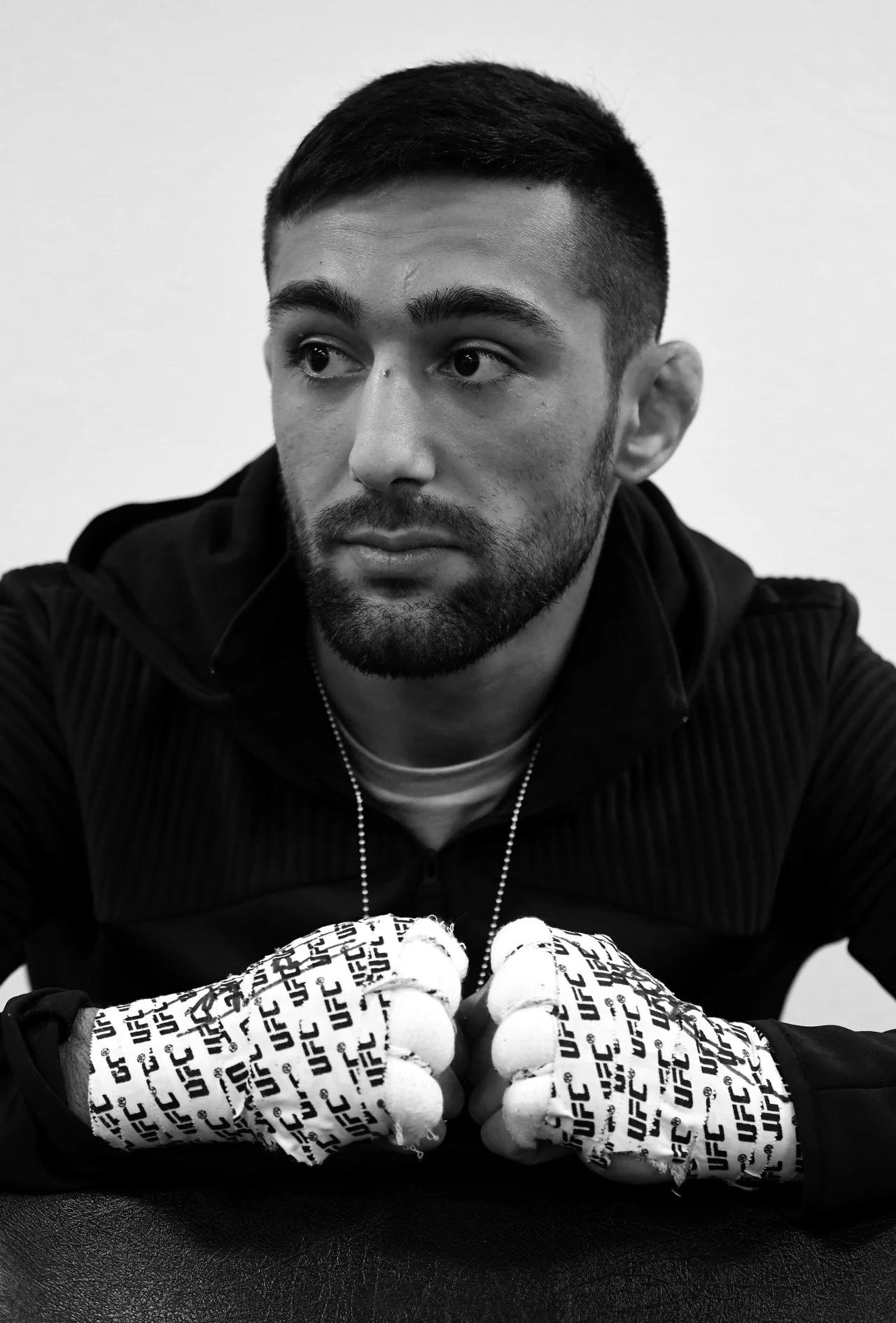 Suman Mokhtarian Młody mężczyzna z zarostem siedzi z lekko opuszczoną głową, ubraną w czarną bluzę, na dłoniach ma owijki z logo UFC.