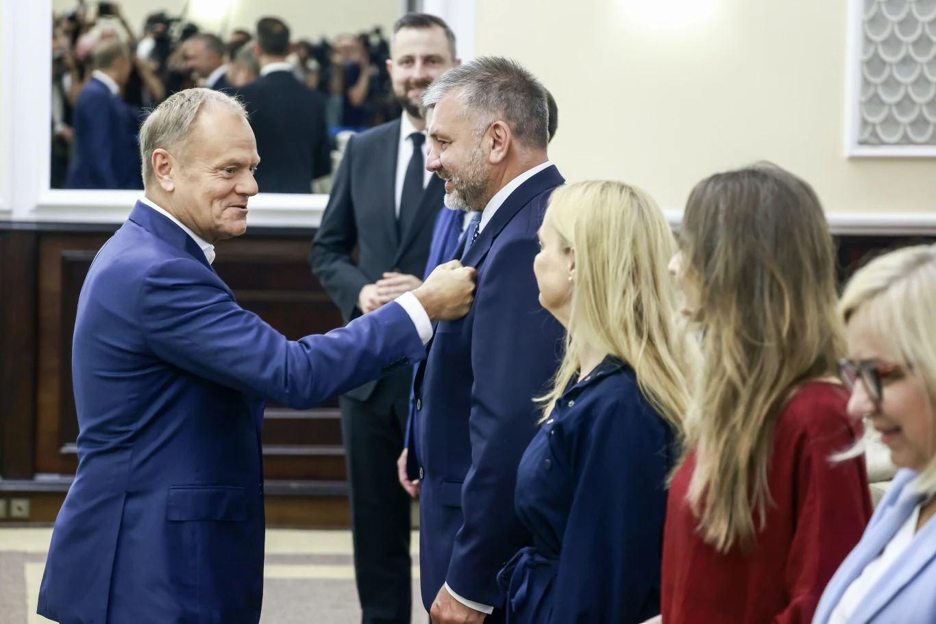 Mężczyzna w garniturze przypina odznaczenie innemu mężczyźnie, a obok stoją trzy kobiety w eleganckich strojach, sala urzędowa z lustrami i drewnianymi panelami, w tle widoczni fotografowie i obserwatorzy.