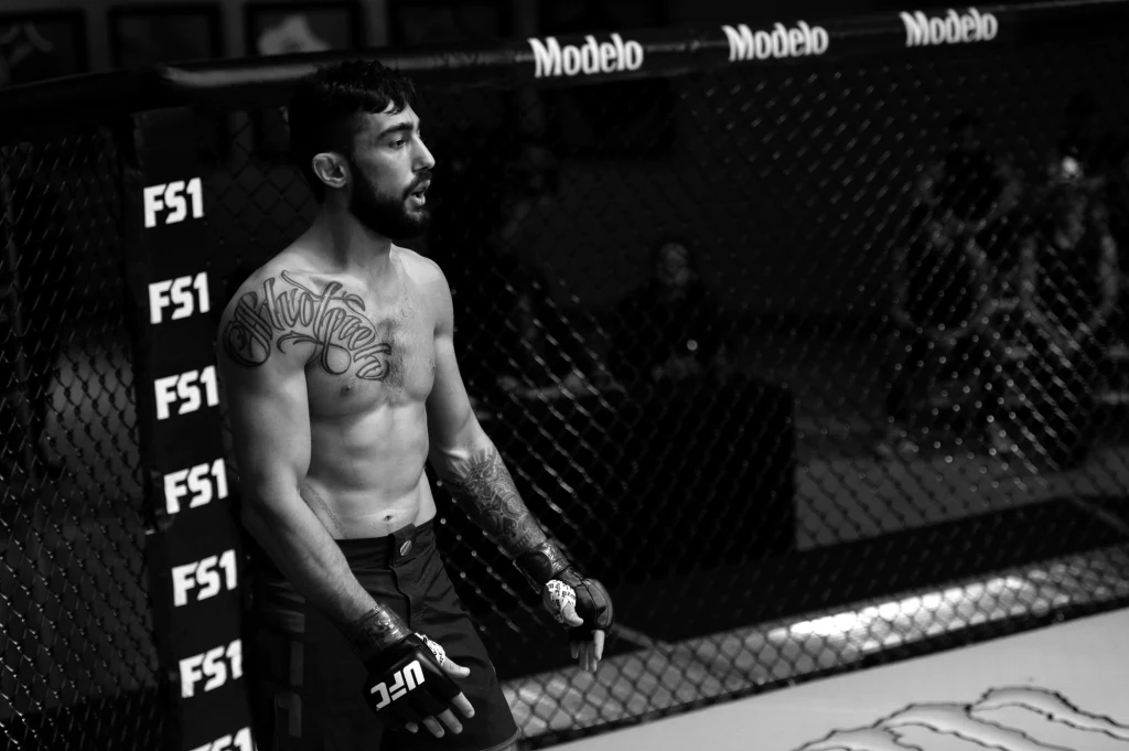 Suman Mokhtarian Zawodnik MMA stojący w klatce ringu, bez koszulki, z widocznymi tatuażami na ramieniu i klatce piersiowej, ręce owinięte taśmą ochronną, w tle siatka oraz logo sponsorów.