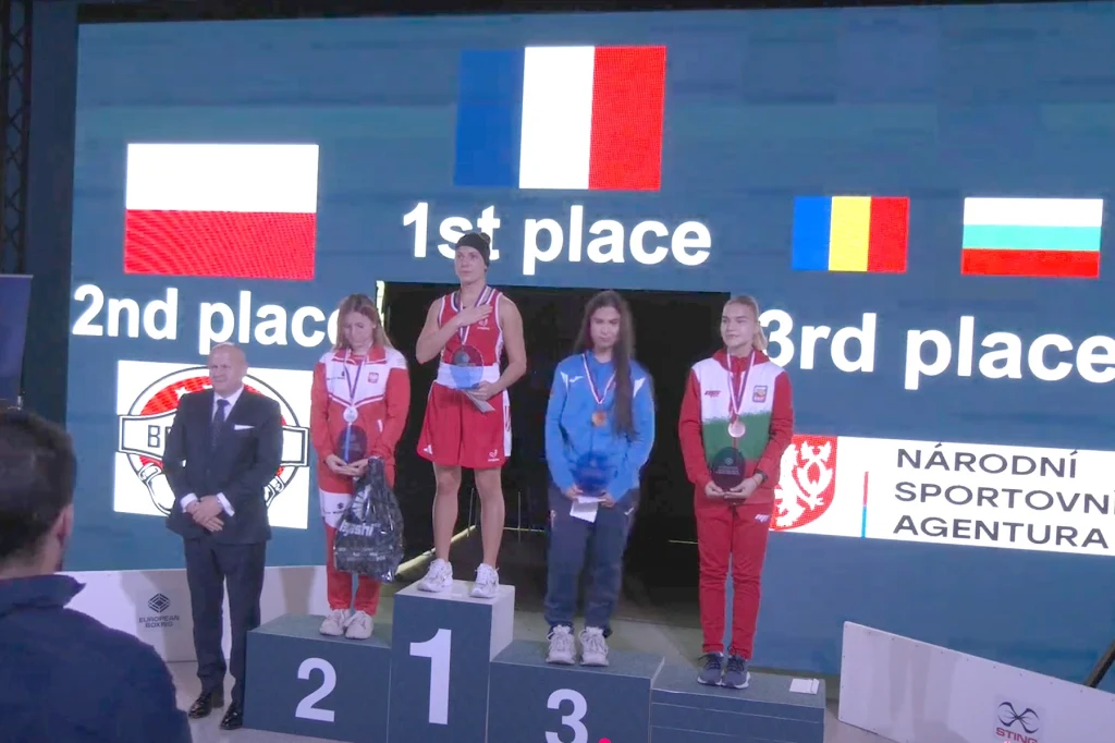 Trzy zawodniczki stoją na podium z medalami, za nimi ekran z flagami państw i miejscami na podium, obok znajduje się oficjalny przedstawiciel zawodów.