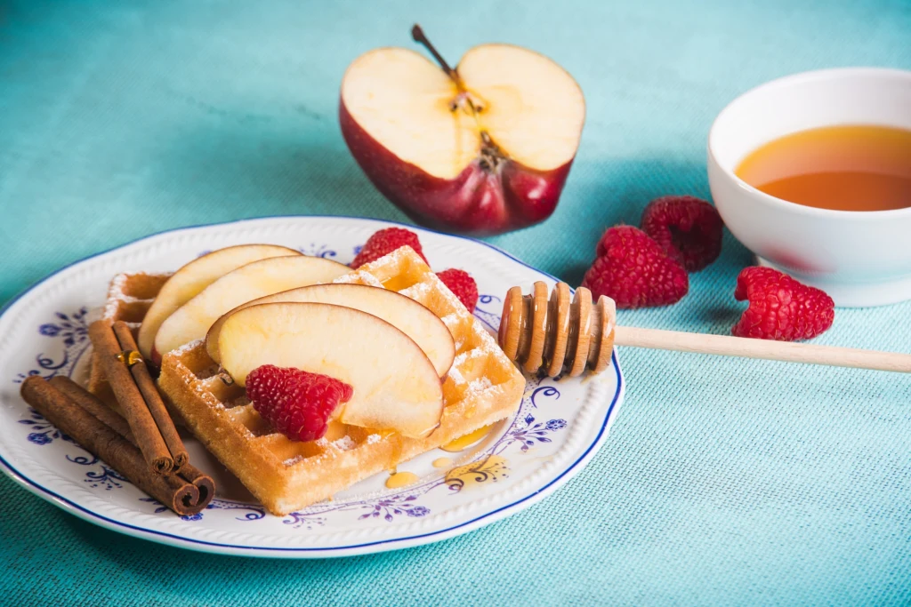 Gofry z dodatkiem jabłek i cynamonu będą nie tylko smaczne ale i bardziej sycące Gofry z plasterkami świeżego jabłka i malinami na porcelanowym talerzu, ozdobione laskami cynamonu i polane miodem, w tle połówka jabłka, kilka malin i miseczka z miodem.