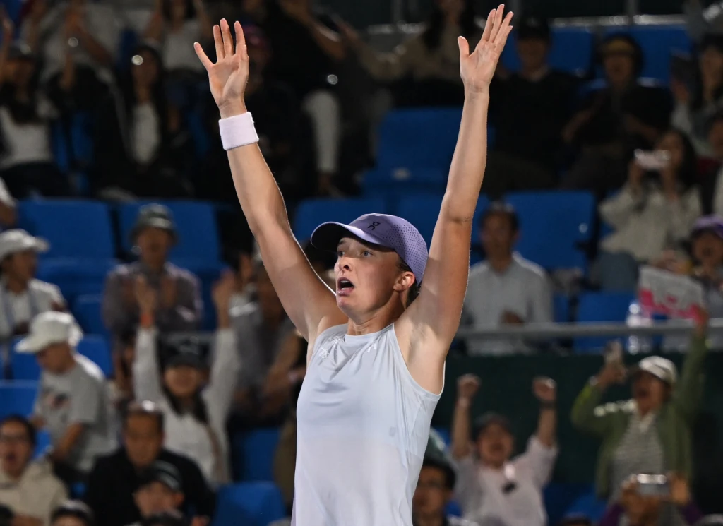 Iga Świątek Zawodniczka tenisowa unosi ręce w geście triumfu na korcie, wokół niej widać wiwatującą publiczność na trybunach.