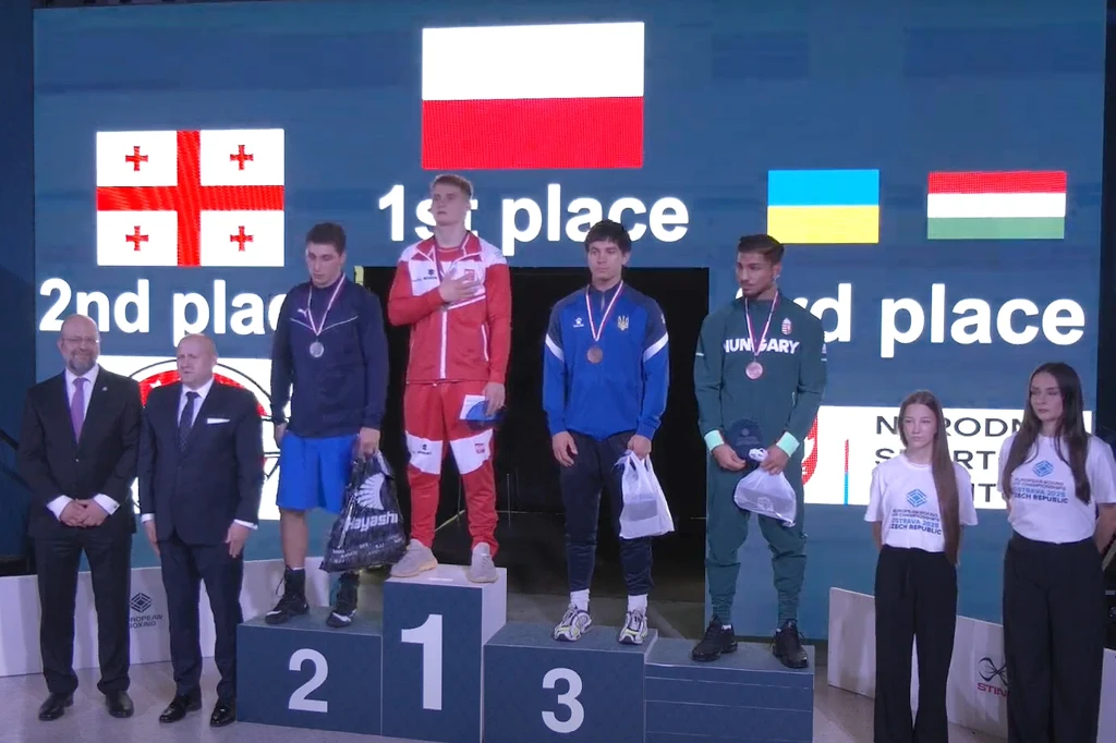 Trzech młodych sportowców stoi na podium, trzymając medale, w tle ekrany z flagami Polski, Gruzji, Ukrainy oraz Węgier, obok nich widoczni są sędziowie oraz hostessy.