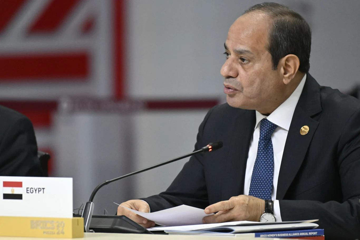 Prezydent Egiptu Abdel Fattah el-Sisi Mężczyzna w garniturze i krawacie siedzi przy stole konferencyjnym z mikrofonem, przed nim znajduje się tabliczka z napisem Egypt oraz flaga tego kraju, trzyma dokumenty, w tle widoczne elementy sali konferencyjnej.