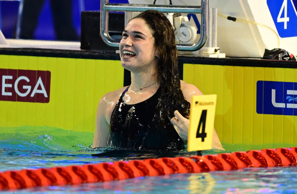 Benedetta Pilato Zadowolona młoda kobieta w stroju kąpielowym stoi w basenie przy torze numer 4 po zakończonym wyścigu, uśmiecha się i unosi dłonią gest zwycięstwa, w tle widoczne oznaczenia basenu i tablice wyników.