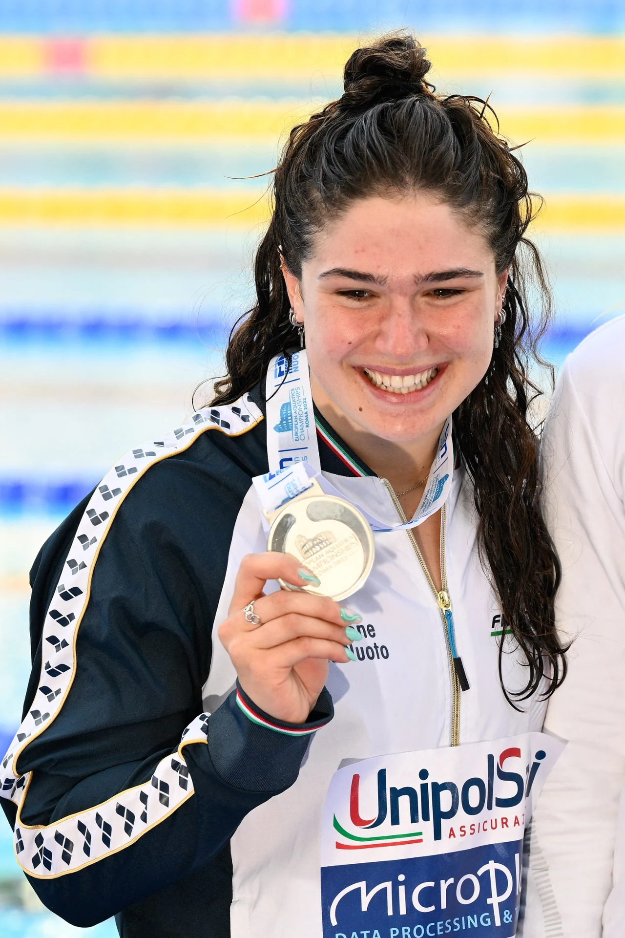 Benedetta Pilato Młoda kobieta w sportowym stroju z mokrymi włosami szeroko się uśmiecha, trzymając srebrny medal na tle basenu olimpijskiego.