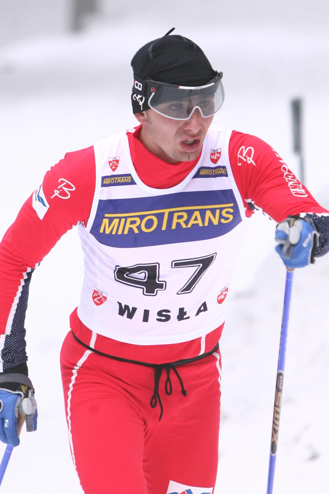 Zawodnik w czerwonym stroju narciarskim bierze udział w biegu narciarskim, trzymając kijki i nosząc numer startowy 47 oraz gogle ochronne, na tle ośnieżonego krajobrazu.