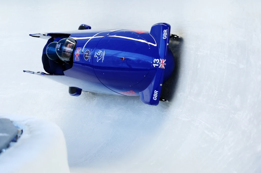 Arran Gulliver podczas rywalizacji Niebieski bobslej z oznaczeniem GBR i numerem 13 ślizga się po lodowym torze podczas zawodów zimowych, prowadzi go sportowiec w kasku z flagą Wielkiej Brytanii.