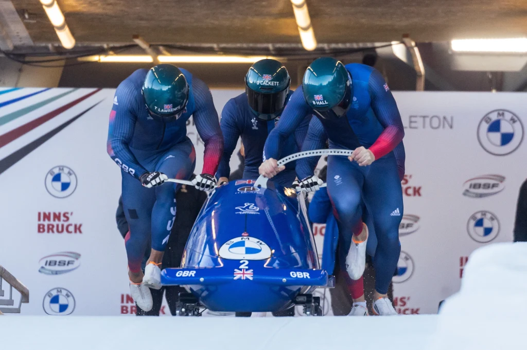 Brad All, Arran Gulliver, Taylor Lawrence i Greg Cackett Czterech sportowców w kombinezonach startuje z wózkiem bobslejowym na torze, wszyscy koncentrują się na pchaniu pojazdu, tło zdobią logotypy sponsorów i oznaczenia zawodów.