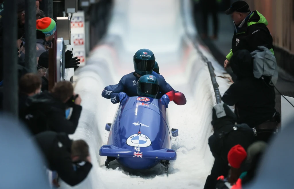 Great Britain's Brad All, Arran Gulliver, Taylor Lawrence i Greg Cackett Dwóch sportowców w niebieskim bobsleju z flagą Wielkiej Brytanii, zjeżdżających po torze lodowym, otoczonych przez kibiców i sędziów.