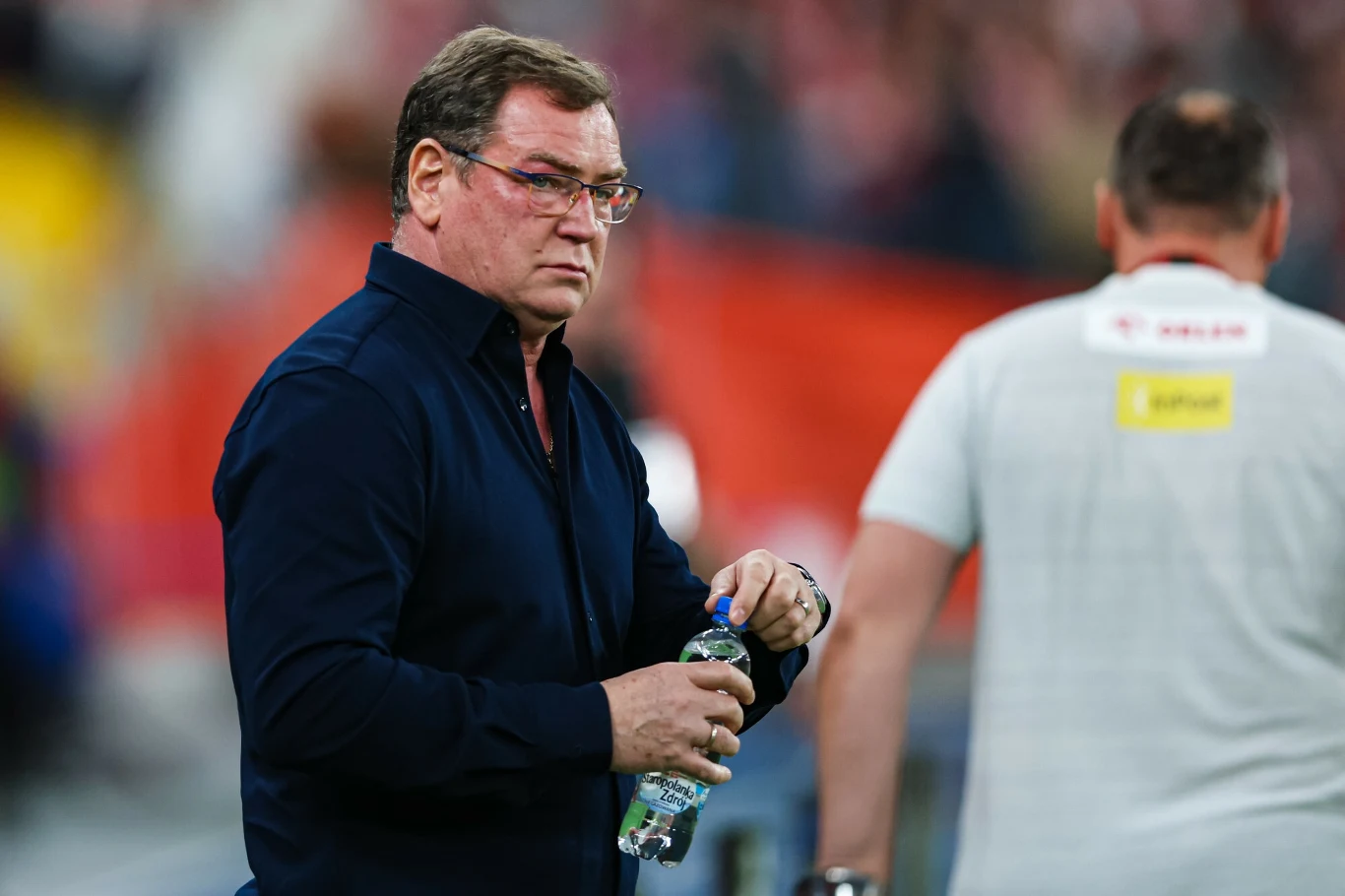 Jan Urban podczas meczu reprezentacji Polski Mężczyzna w ciemnej koszuli trzymający butelkę wody na tle rozmytego stadionu, w tle inna osoba w białej koszulce z odwróconymi plecami.