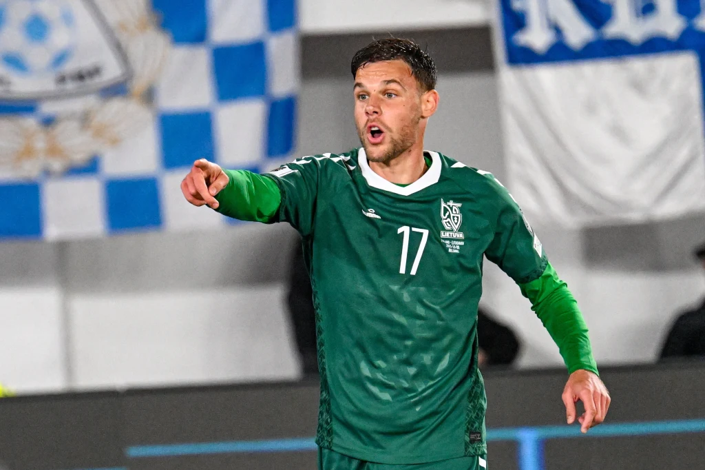 Piłkarz ubrany w zielony strój z numerem 17 wskazuje ręką na coś poza kadrem, w tle widoczne nieostre flagi kibiców.