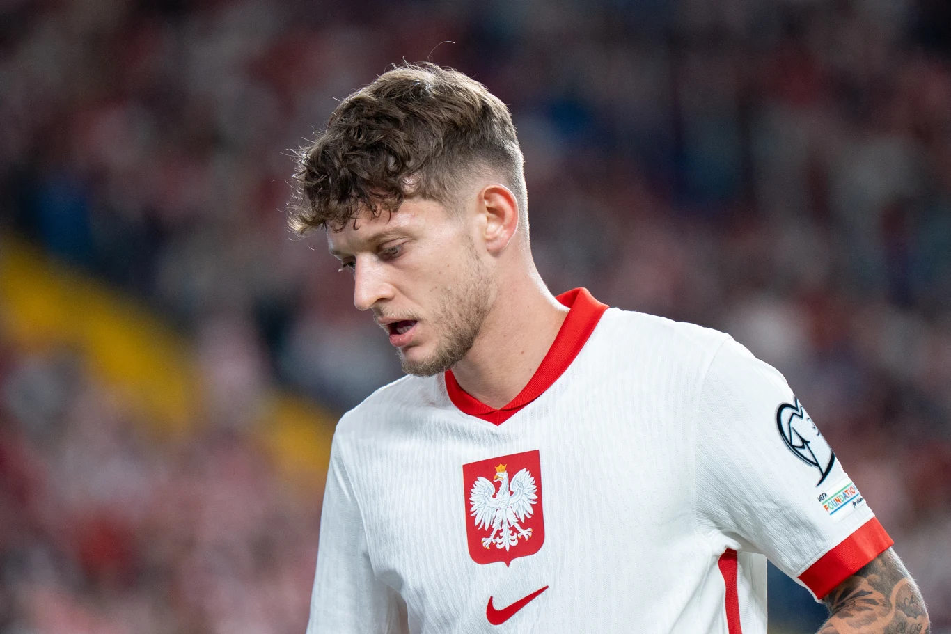 Piłkarz w białej koszulce reprezentacji Polski z godłem oraz czerwonym kołnierzem, na tle rozmytego stadionu wypełnionego kibicami.