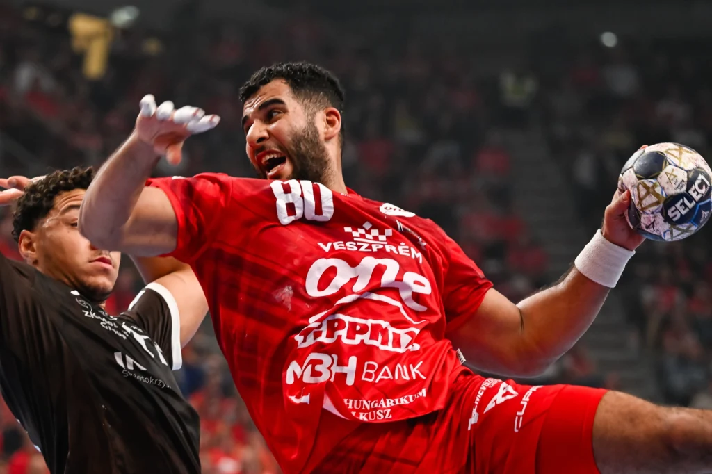 Zawodnik w czerwonym stroju podczas dynamicznej akcji na meczu piłki ręcznej, trzyma piłkę w prawej dłoni i jest skierowany w stronę bramki, obok widoczny zawodnik drużyny przeciwnej w czarnej koszulce.