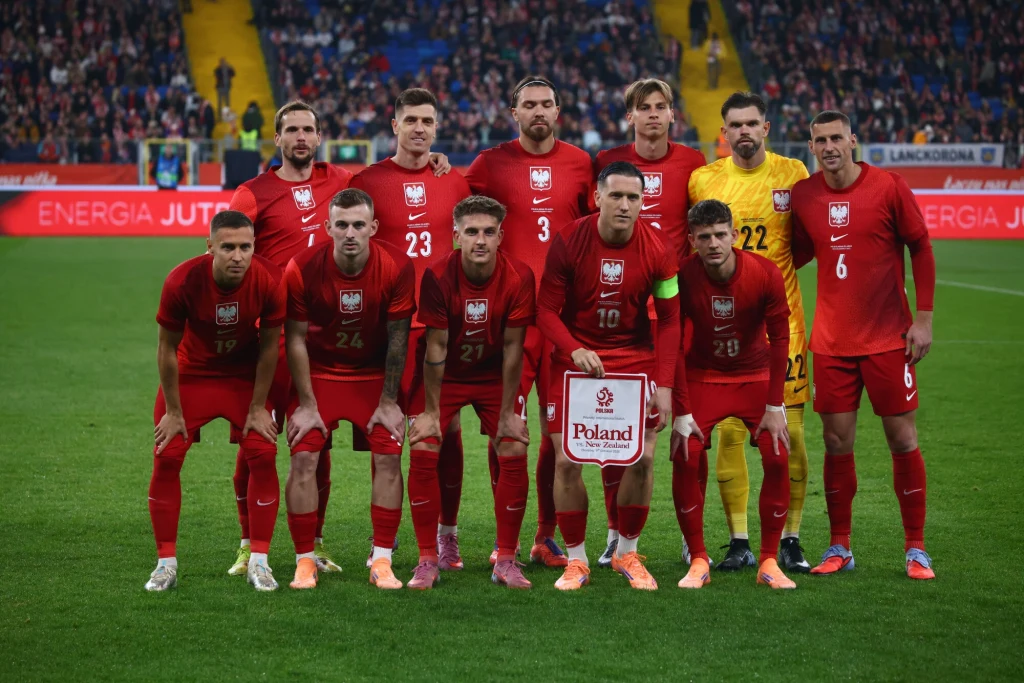 Reprezentacja Polski w meczu z Nową Zelandią (9.10.2025) Polska reprezentacja piłkarska ustawiona w dwóch rzędach przed rozpoczęciem meczu, piłkarze ubrani w czerwone stroje, w tle trybuny pełne kibiców.