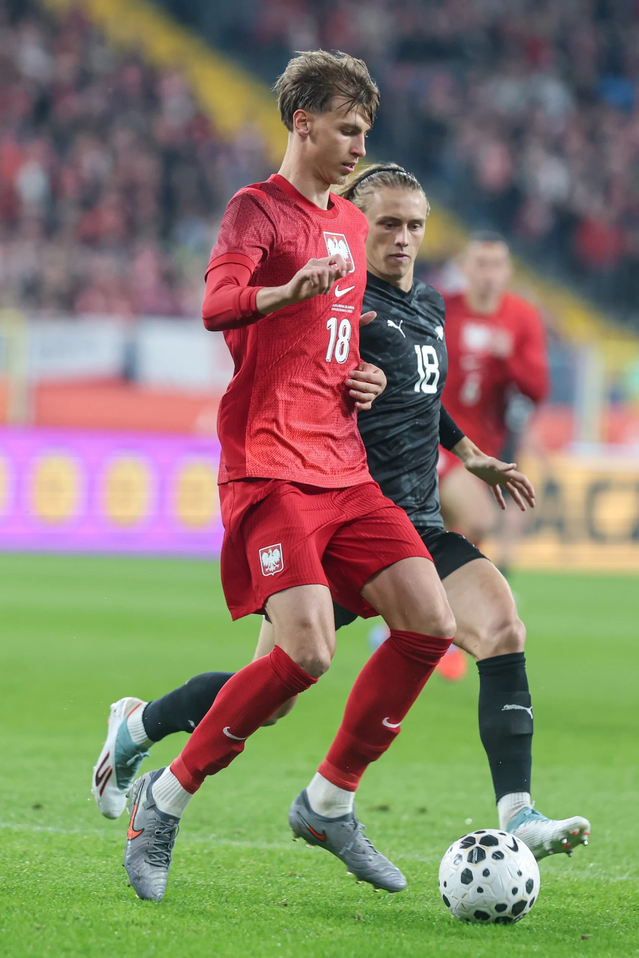 Dwóch piłkarzy rywalizujących o piłkę podczas meczu, pierwszy z nich w czerwonym stroju reprezentacji Polski, drugi w czarnym stroju drużyny przeciwnej, dynamiczna akcja na murawie stadionu