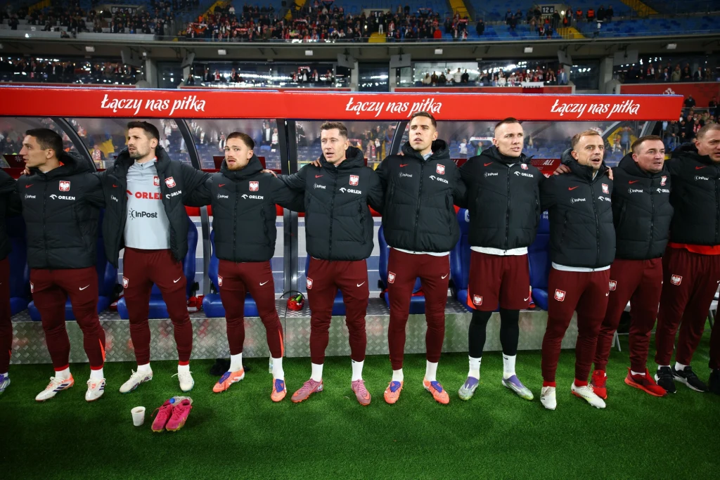 Grupa piłkarzy w sportowych strojach, stojących ramię w ramię na stadionie, z widoczną ławką rezerwowych i napisem 'Łączy nas piłka' w tle, na trybunach liczni kibice.