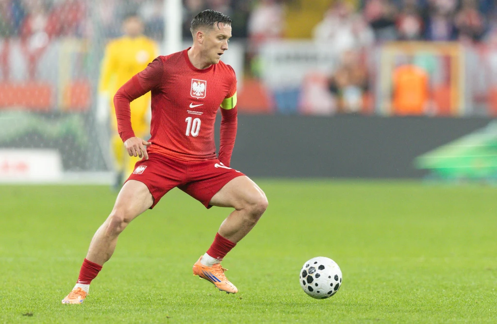 Piłkarz w czerwonym stroju reprezentacji Polski z numerem 10 kontroluje piłkę na murawie podczas meczu, w tle widać innych zawodników oraz rozmyte trybuny.