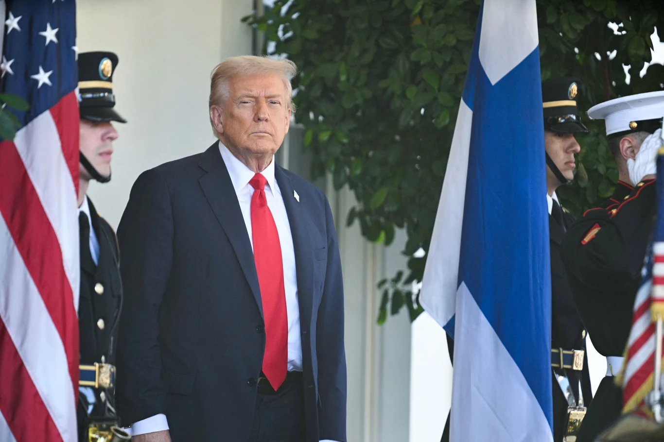 Donald Trump sugeruje możliwość wyrzucenia Hiszpanii z NATO Mężczyzna w garniturze i czerwonym krawacie stoi pomiędzy żołnierzami w mundurach, obok flag Stanów Zjednoczonych oraz Finlandii.