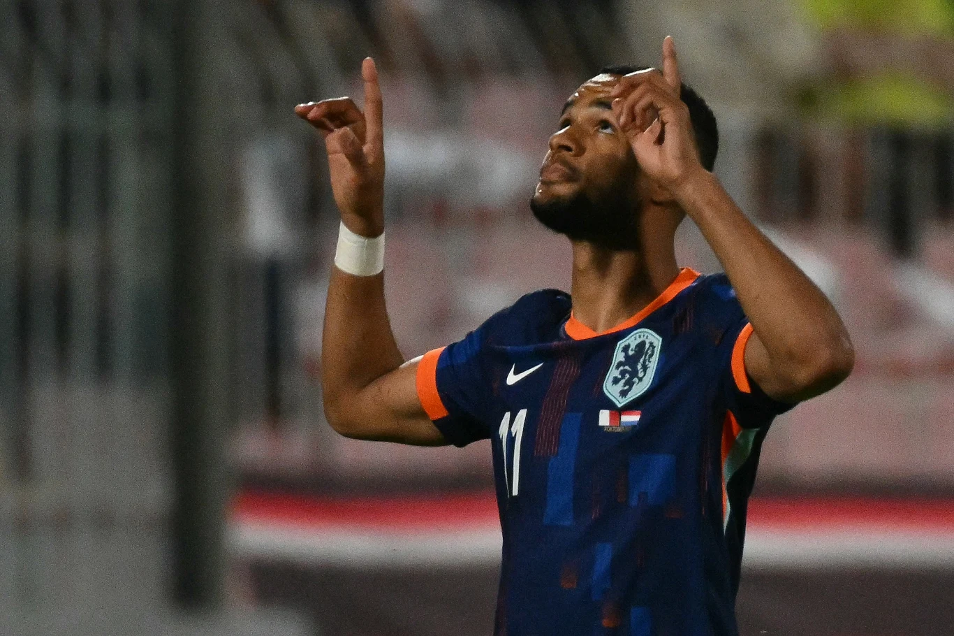 Piłkarz w granatowo-pomarańczowej koszulce z numerem 11 i herbem reprezentacji Holandii unosi ręce do góry, w tle rozmyte trybuny stadionu.