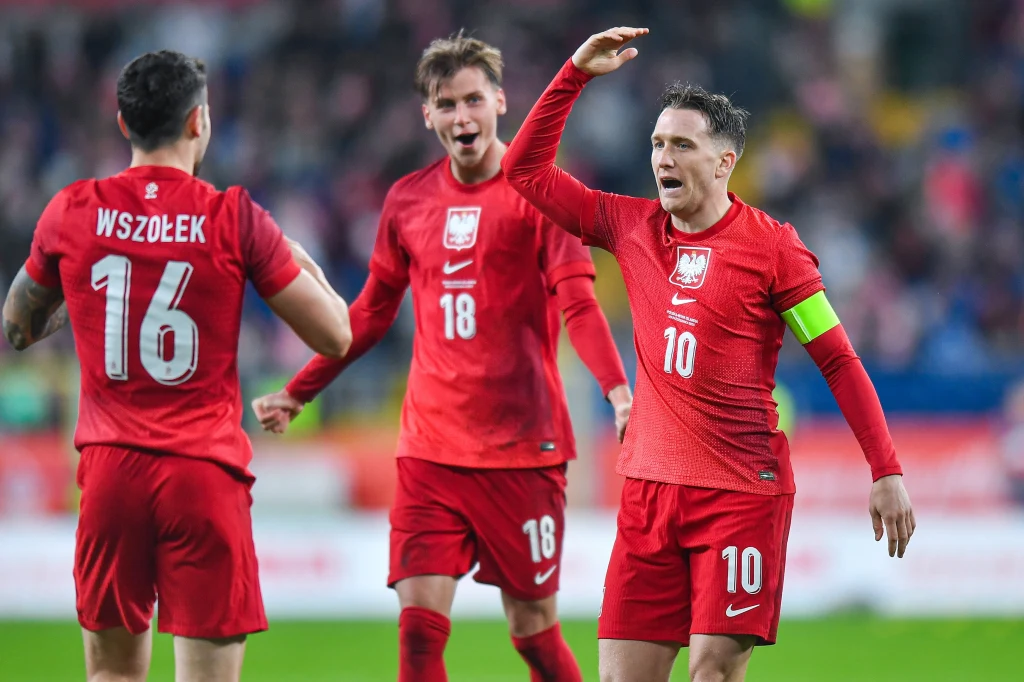 Do triumfu poprowadził "Biało-Czerwonych" kapitan Piotr Zieliński. Kapitalne uderzenie z 49. minuty rozstrzygnęło losy spotkania Trzech polskich piłkarzy w czerwonych strojach narodowych na boisku podczas meczu, jeden zawodnik nosi opaskę kapitana i gestykuluje, w tle rozmyta publiczność.