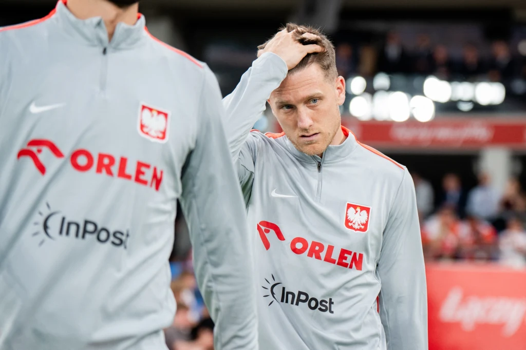 Piłkarz reprezentacji Polski w szarej bluzie dresowej z logotypami sponsorów i godłem Polski na piersi, z ręką na głowie, stoi na tle stadionu obok innego członka drużyny.