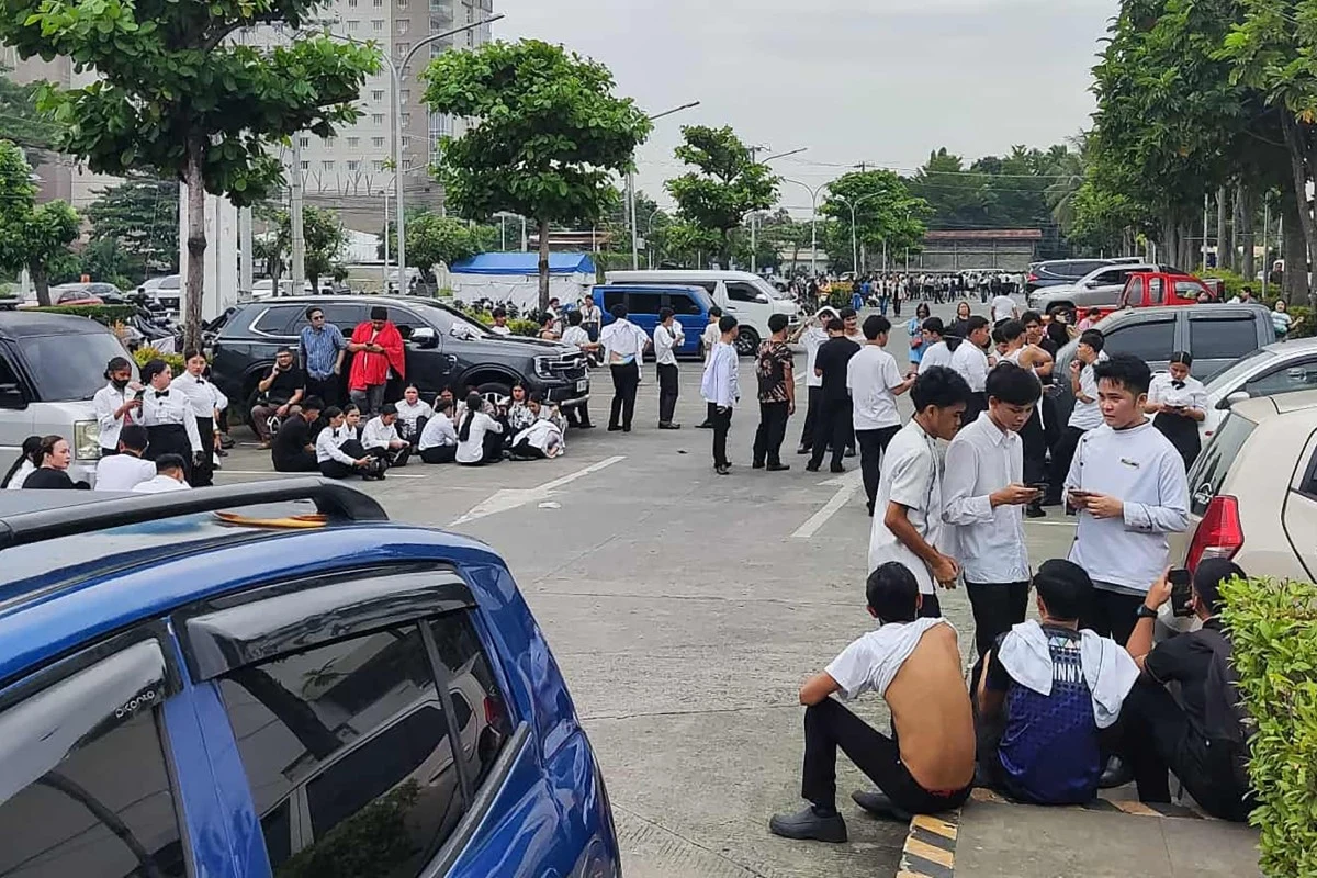 Pracownicy centrum handlowego w Davao City (Mindanao) ewakuowani po trzęsieniu ziemi Grupa młodych osób ubranych w formalne stroje zgromadzona na parkingu w miejskiej scenerii, niektórzy siedzą na chodniku, inni rozmawiają na stojąco, w tle samochody oraz drzewa.