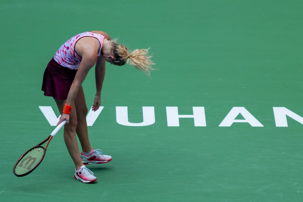 Zawodniczka tenisowa pochylona ze zmęczenia lub rozczarowania podczas turnieju na zielonym korcie z napisem WUHAN. Ubrana w sportowy strój, trzyma rakietę opartą o ziemię.