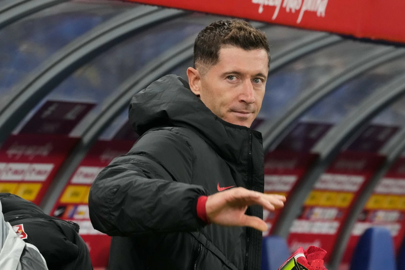 Piłkarz w czarnej kurtce gestykulujący dłonią, stojący na tle ławki rezerwowych stadionu sportowego.