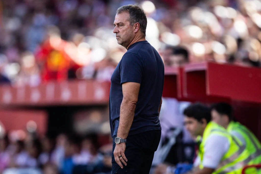 Hansi Flick Mężczyzna w ciemnej koszulce stoi na tle trybun, z poważnym wyrazem twarzy, w tle rozmyte postacie kibiców i pracowników obsługi ubrane w żółte kamizelki