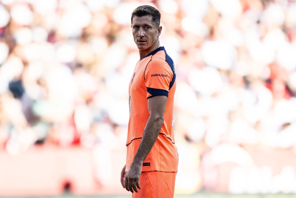 Robert Lewandowski Piłkarz w pomarańczowym stroju sportowym stoi na boisku podczas meczu, wokół tłum kibiców w rozmytym tle, na jego twarzy skupiony wyraz.