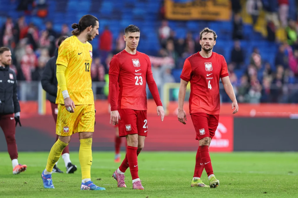 Krzysztof Piątek (w środku) Trzech piłkarzy reprezentacji Polski w oficjalnych strojach sportowych, dwóch ubranych w czerwone koszulki i jeden w żółtym stroju bramkarskim, idzie po murawie boiska, w tle widoczna rozmyta publiczność oraz sektor kibiców.
