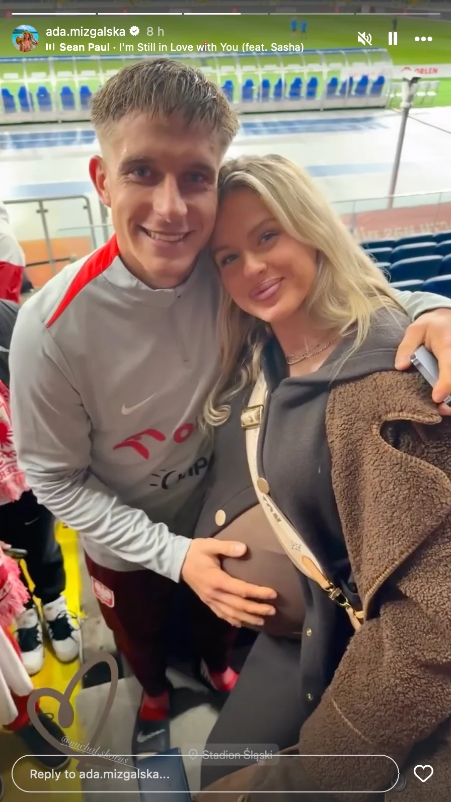 Ada Mizgalska wspierała Michała Skórasia podczas meczu z Nową Zelandią Para obejmująca się na trybunach stadionu, mężczyzna trzyma rękę na brzuchu ciężarnej kobiety, oboje uśmiechnięci, w tle widoczne puste miejsca siedzące i fragment bieżni sportowej.