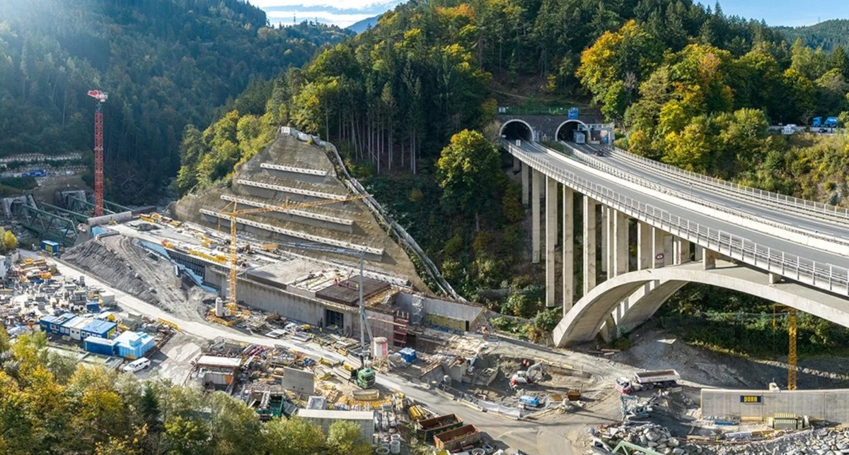 Tunel Bazowy Brenner najdłuższy na świecie. Powstaje pod Alpami. Nowoczesny wiadukt oraz tunel w górskim otoczeniu, sąsiadujące z rozległym placem budowy, na którym pracuje ciężki sprzęt. Zalesione zbocza oraz roślinność kontrastują z infrastrukturą drogową i prowadzonymi robotami.