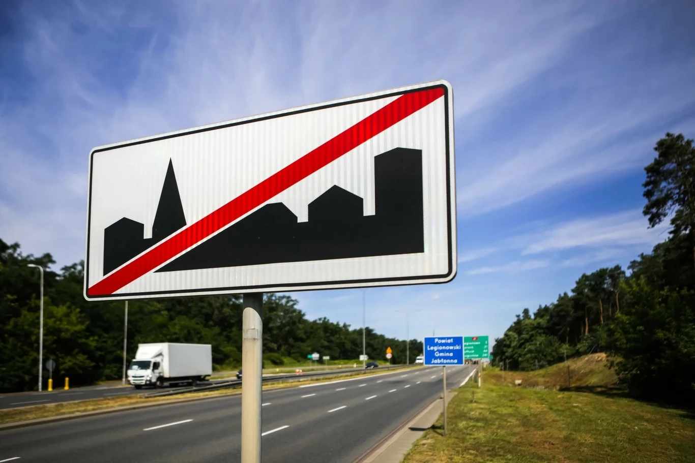 Za znakiem D-43 nowy mandat grozi nie kierowcom, ale pieszym Znak drogowy oznaczający koniec obszaru zabudowanego ustawiony przy dwupasmowej, asfaltowej drodze otoczonej zielenią i lasem, w tle tablice kierunkowe oraz ciężarówka jadąca w stronę miasta.