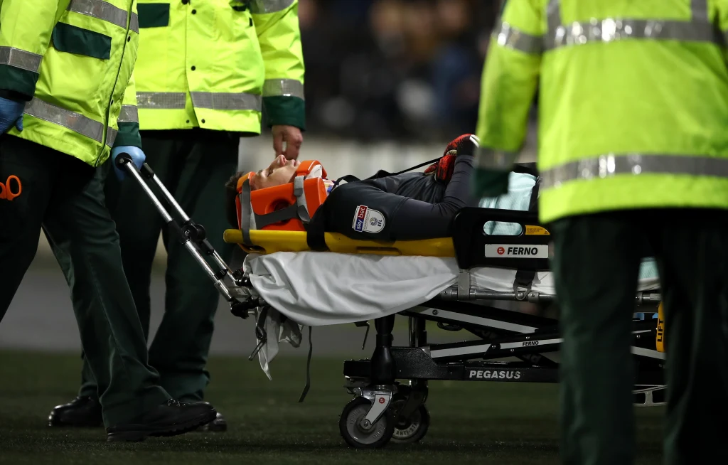 Ratownicy medyczni przewożą osobę leżącą na noszach, otoczoną przez ratowników w odblaskowych kurtkach, w tle nieostre postaci i ciemne otoczenie.