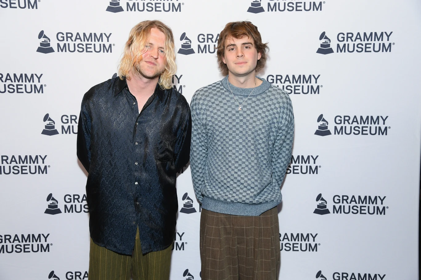 Zespół Royel Otis został headlinerem pierwszego dnia Inside Seaside Dwaj młodzi mężczyźni stoją obok siebie na tle ścianki z logo Grammy Museum, obaj w swobodnych stylizacjach, jeden ubrany w czarną koszulę i zielone spodnie, drugi w szary sweter i brązowe spodnie, obaj patrzą prosto do obiektywu