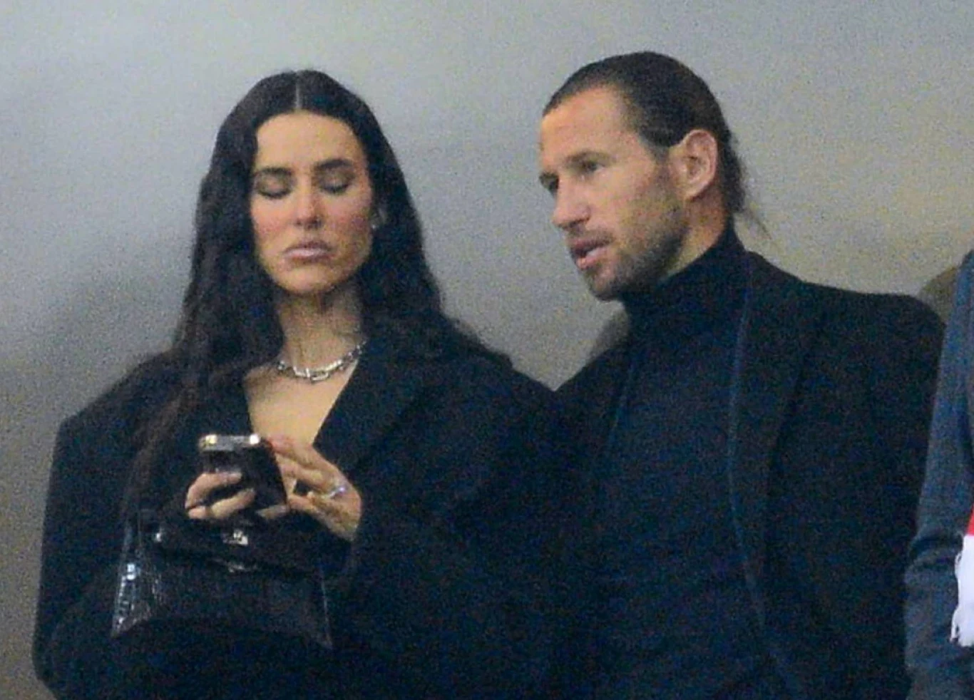 Celia Jaunat i Grzegorz Krychowiak Dwie osoby stoją obok siebie, kobieta skupiona na telefonie, mężczyzna zwrócony w jej stronę, oboje ubrani elegancko, na tle neutralnym.