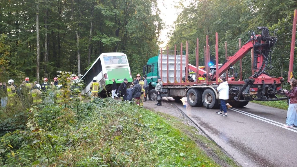 Autobus znajdujący się w rowie przy lesie oraz ciężarówka z załadunkiem ustawiona na poboczu, wokół zgromadzeni strażacy i służby ratownicze zabezpieczające miejsce zdarzenia, szosa zablokowana częściowo przez pojazdy ratunkowe.