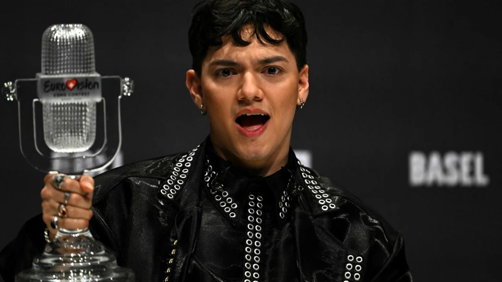 Osoba w ciemnym stroju trzymająca statuetkę mikrofonu z napisem Eurovision, wyrażająca emocje na tle ciemnego, oficjalnego tła z napisem Basel.