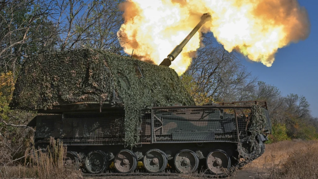 Armatohaubica Krab w Ukrainie Samobieżna armatohaubica oddająca strzał, częściowo pokryta siatką maskującą, otoczona suchą roślinnością, w tle drzewa bez liści i intensywny błysk powstały w momencie wystrzału.