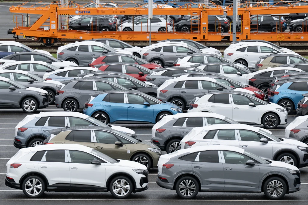 Szeregi nowych samochodów osobowych zaparkowanych na dużym placu, różne modele i kolory, w tle widoczny pociąg towarowy przewożący kolejne pojazdy na lawetach.