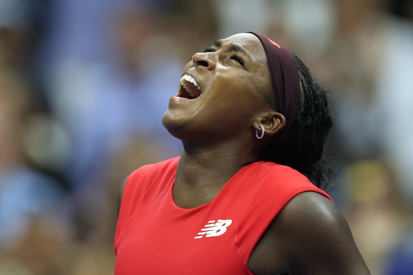 Tenisistka w czerwonym stroju z opaską na głowie wyraża silne emocje, podnosząc głowę i otwierając usta szeroko, w tle rozmyta publiczność.