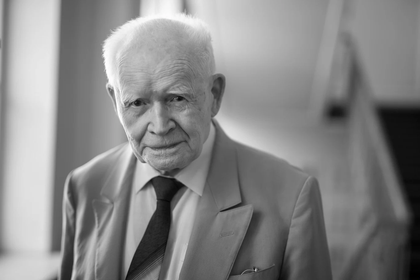 Prof. Adam Strzembosz nie żyje Starszy mężczyzna w garniturze i krawacie patrzy bezpośrednio w obiektyw, stoi w jasnym, rozmytym wnętrzu, wyrażając spokojną i pełną życzliwości postawę.