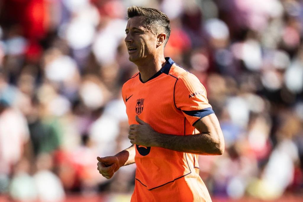 Robert Lewandowski Piłkarz w pomarańczowym stroju sportowym biegnący na boisku podczas meczu, w tle rozmyte sylwetki innych osób i widownia.