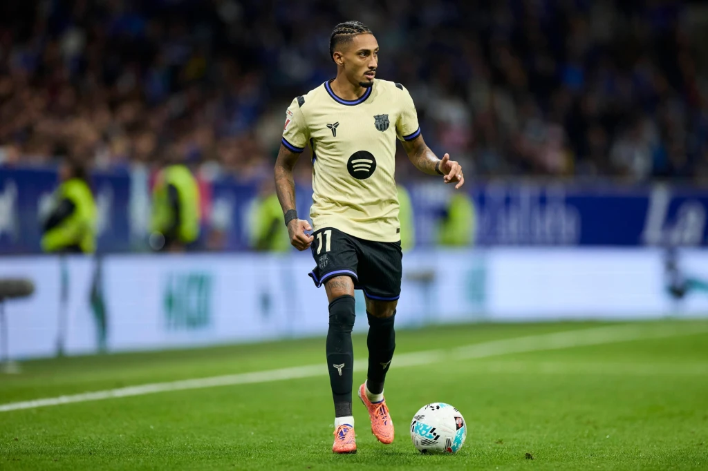 Raphinha Piłkarz w beżowej koszulce drużyny FC Barcelona prowadzi piłkę nożną po boisku podczas meczu, w tle widoczna rozmyta publiczność oraz elementy stadionu.