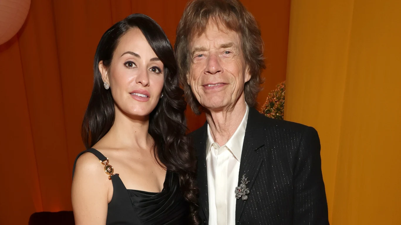 Mick Jagger (The Rolling Stones) i jego partnerka Melanie Hamrick Para elegancko ubranych osób pozuje razem na tle pomarańczowego oświetlenia, oboje uśmiechają się delikatnie do aparatu.