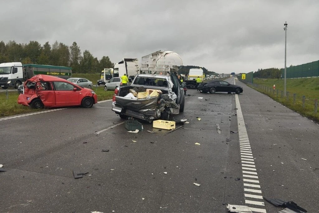 Karambol na A1 Rozbite samochody rozrzucone na autostradzie, widoczne uszkodzenia wielu pojazdów, pojazdy służb ratowniczych w tle, mokra nawierzchnia i pochmurne niebo, elementy wyposażenia samochodów rozrzucone wokół miejsca zdarzenia.