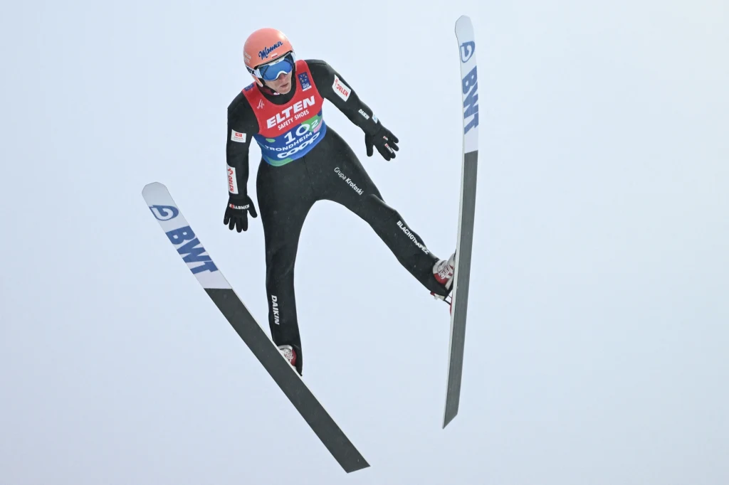 Dawid Kubacki na poprzednich zimowych igrzyskach olimpijskich zdobył brązowy medal Skoczek narciarski w locie z wyciągniętymi rękami i nartami szeroko rozstawionymi, w pełnym stroju sportowym oraz kasku i goglach, na tle jasnego nieba.