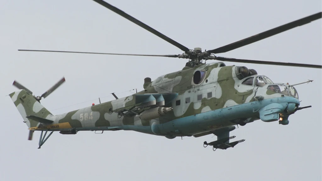 Polski śmigłowiec szturmowy Mil Mi-24 Śmigłowiec bojowy w kamuflażu z widocznym uzbrojeniem, lecący na tle jasnego nieba.